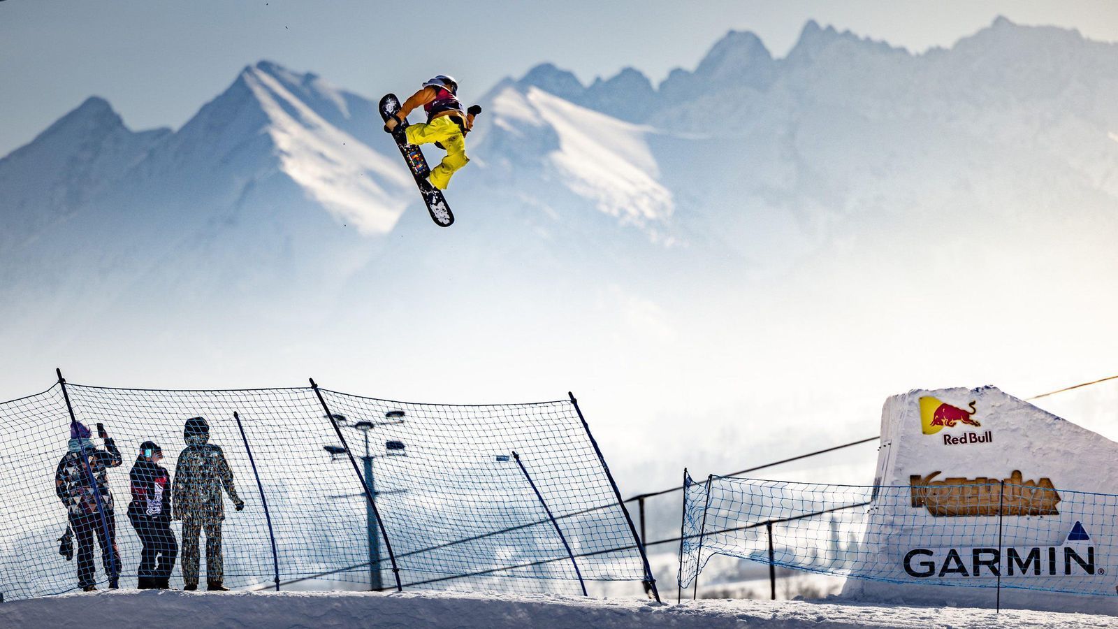 Snowboardzista w żółtych spodniach w locie nad siatką, w tle ośnieżone szczyty